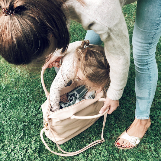 Mutter und Kleinkind blicken in eine beigefarbene Handtasche mit grauen Lilibell Organizer-Einsatz für Babyutensilien auf einer Wiese.