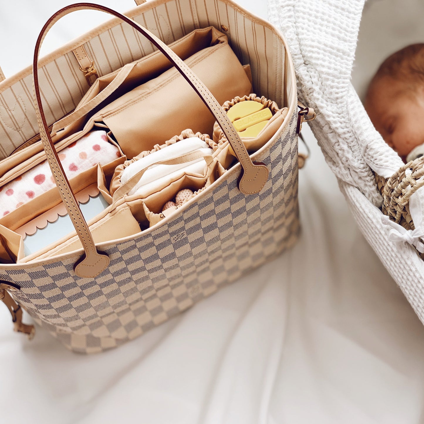 Eine stilvolle Wickeltasche Louis Vuitton Neverfull MM mit einem praktischen Organizer-Einsatz, gefüllt mit Baby-Essentials wie Windeln, einer Snackbox, einer gehäkelten Rassel und einem Schnullerhalter. Die Tasche liegt neben einem schlafenden Neugeborenen in einem weichen Babynest. Der Organizer Classic von Lilibell, sorgt für Ordnung und schnellen Zugriff – ideal für stilbewusste Eltern, die unterwegs gut organisiert sein möchten.