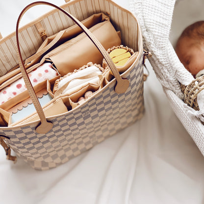 Eine stilvolle Wickeltasche Louis Vuitton Neverfull MM mit einem praktischen Organizer-Einsatz, gefüllt mit Baby-Essentials wie Windeln, einer Snackbox, einer gehäkelten Rassel und einem Schnullerhalter. Die Tasche liegt neben einem schlafenden Neugeborenen in einem weichen Babynest. Der Organizer Classic von Lilibell, sorgt für Ordnung und schnellen Zugriff – ideal für stilbewusste Eltern, die unterwegs gut organisiert sein möchten.