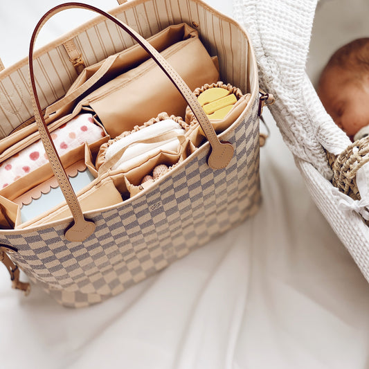 Eine stilvolle Wickeltasche Louis Vuitton Neverfull MM mit einem praktischen Organizer-Einsatz, gefüllt mit Baby-Essentials wie Windeln, einer Snackbox, einer gehäkelten Rassel und einem Schnullerhalter. Die Tasche liegt neben einem schlafenden Neugeborenen in einem weichen Babynest. Der Organizer Classic von Lilibell, sorgt für Ordnung und schnellen Zugriff – ideal für stilbewusste Eltern, die unterwegs gut organisiert sein möchten.
